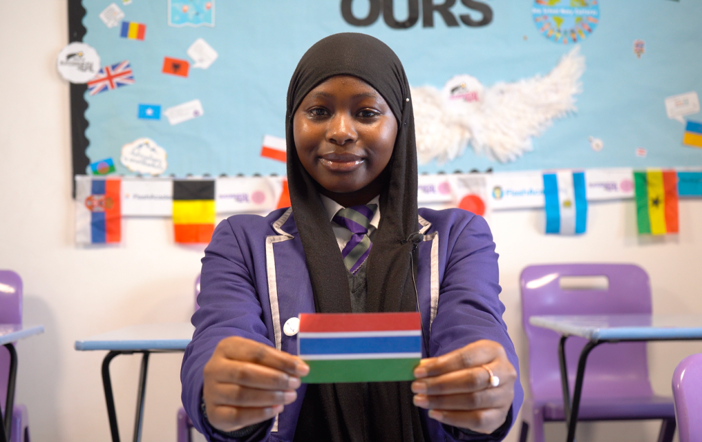 Smiling African female teenager wearing school uniform and hijab holds a small flag of Ghana in front of her.