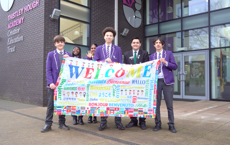 A diverse group of smiling students hold up a colourful welcome sign outside Thistley Hough Academy.