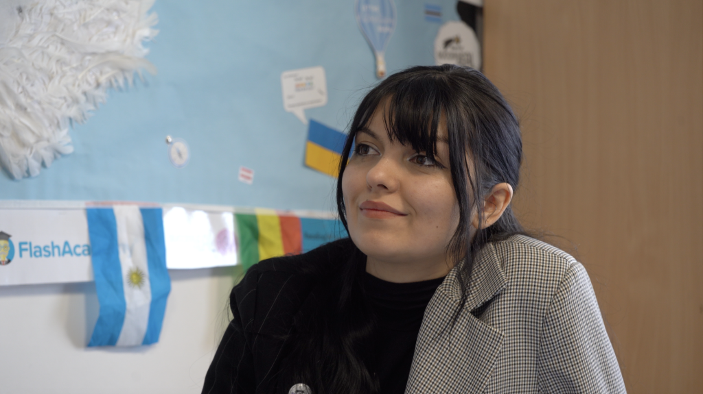 A smiling dark haired female teacher in a suit jacket smiles while sat in the classroom